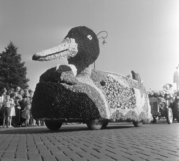 Schwarz-weißes Foto eines Umzugswagens in Form einer Ente, geschmückt mit Blumen, mit Menschen zu seiner Linken und Bäumen im Hintergrund unter einem klaren Himmel.