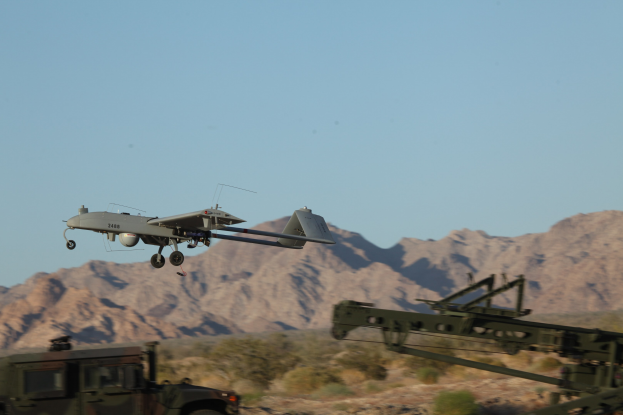 MQ-9 Reaper Unbemanntes Luftfahrzeug fliegt über ein militärisches Fahrzeug in einer Wüstenlandschaft mit Bäumen, Bergen und einem klaren blauen Himmel.