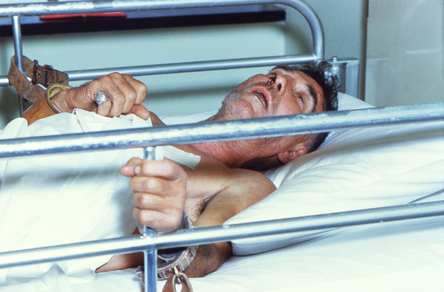 Ein Mann liegt in einem Krankenhausbett mit den Händen auf dem Kopf, der besorgt aussieht mit geschlossenen Augen und geneigtem Körper, umgeben von Metallstangen und einer Wand im Hintergrund.