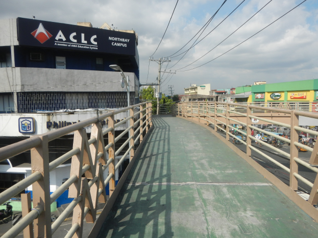A pedestrian bridge with railings and light poles spans over a road with vehicles below, flanked by buildings with signboards, electric poles with wires, trees, and a cloudy sky.