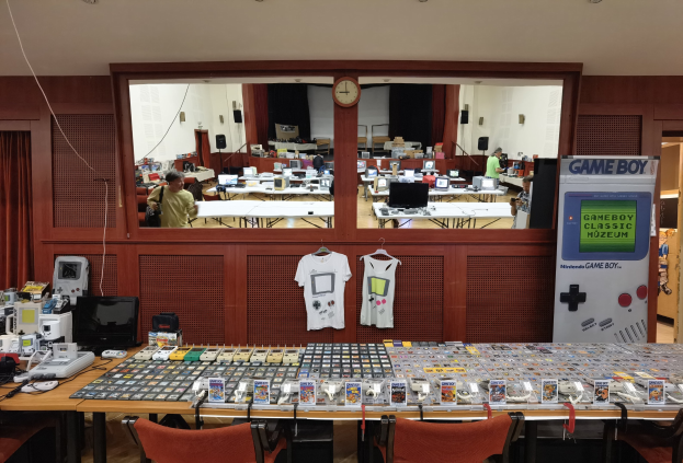 Ein unordentlicher Raum mit Tischen, auf denen Nintendo Game Boy Classic-Artikel, Monitore und T-Shirts präsentiert werden, sowie eine Wanduhr und ein Eingang zu einem weiteren Raum.
