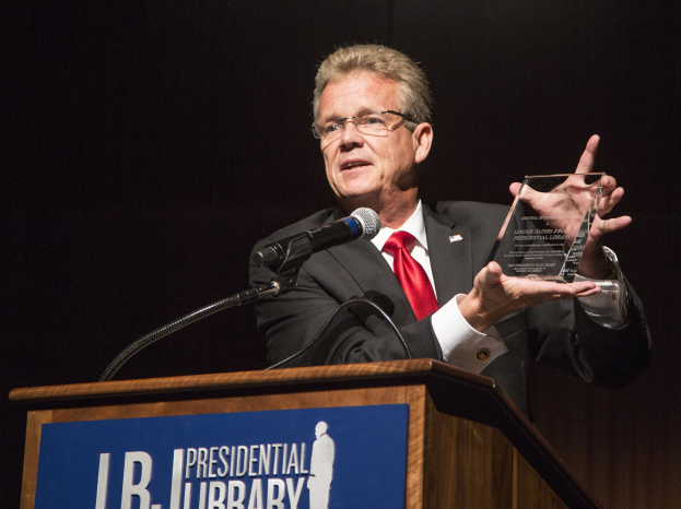Ein Mann im Anzug und Schlips steht hinter einem Rednerpult und hält einen Preis in der Hand, mit einem Mikrofon und einer Tafel daran, trägt eine Brille in einem dunklen Hintergrund bei einer Preisverleihung in einer Bibliothek.