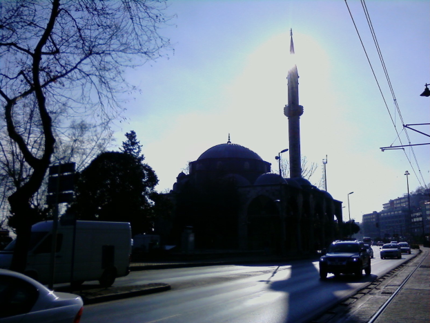 Eine Moschee steht in einer städtischen Straße, umgeben von Gebäuden, Straßeninfrastruktur, Fahrzeugen, Bäumen und einem klaren Himmel.