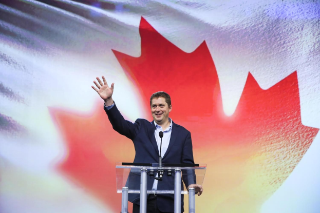 Der kanadische Premierminister Alex Salmond steht an einem Podium mit Mikrofonen, lachend vor einer kanadischen Flagge, mit einem Bildschirm im Hintergrund.