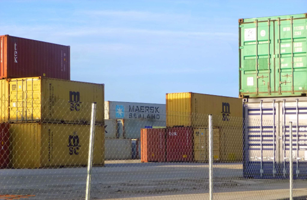 Schiffscontainer in Reihen gestapelt mit Maschendrahtzaun im Vordergrund, Straße darunter und bewölkter Himmel im Hintergrund.