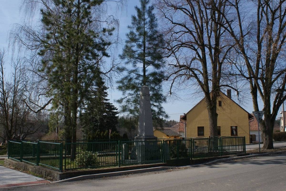 Ein Holocaust-Denkmal steht in der Mitte einer Straße, umgeben von einem Metallzaun, mit Gras, Bäumen, Häusern, einem Straßenmast und einem bewölkten Himmel im Hintergrund.