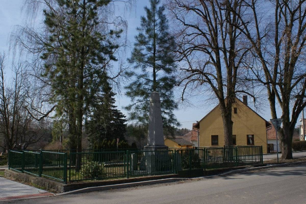Ein Holocaust-Denkmal steht in der Mitte einer Straße, umgeben von einem Metallzaun, mit Gras, Bäumen, Häusern, einem Straßenmast und einem bewölkten Himmel im Hintergrund.