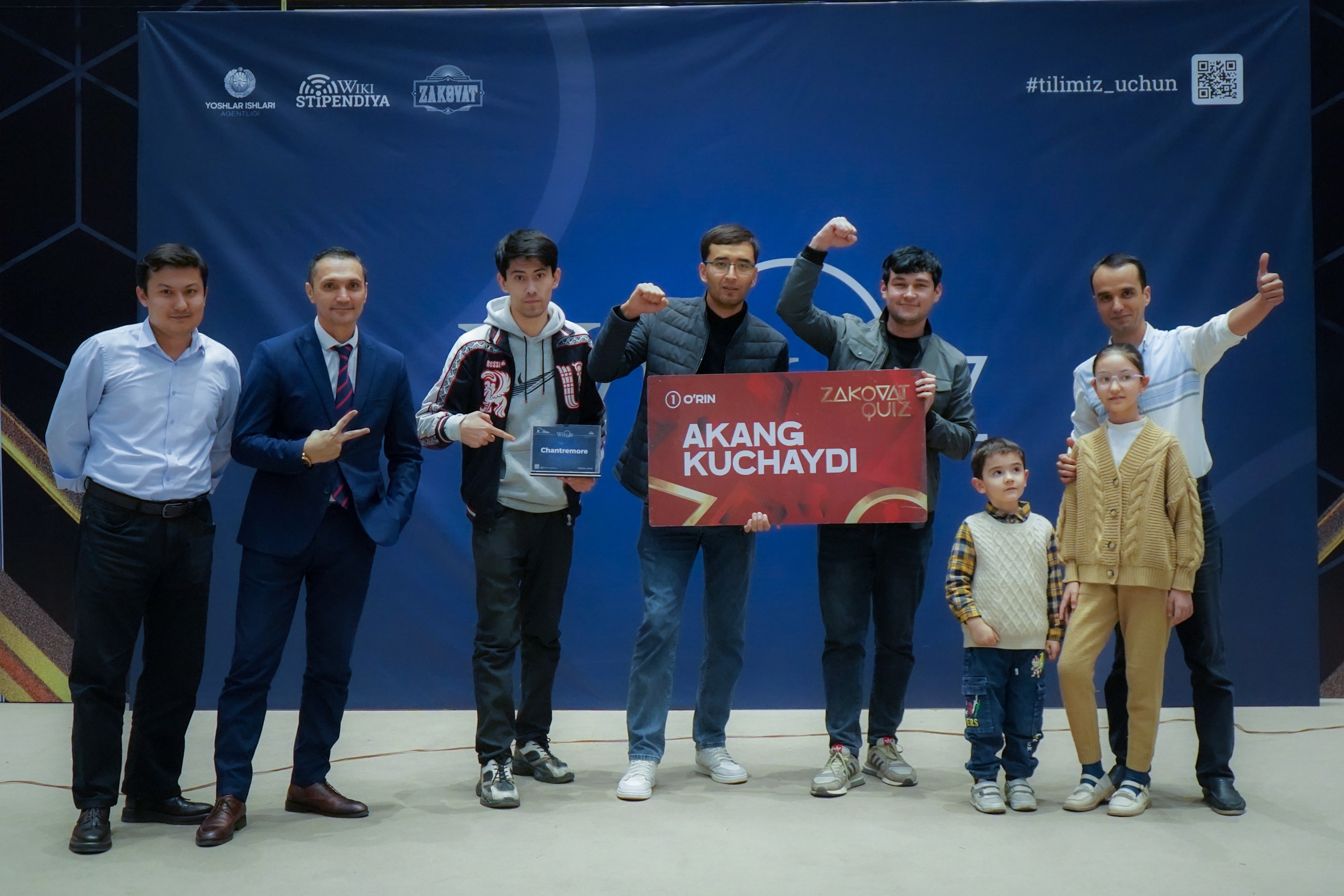 Group of people on a stage holding a board reading "Akang Kuchaydi - Zakovat Quiz" with a banner, barcode, and lights in the background.