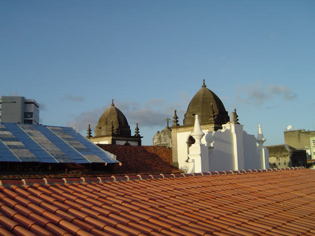 Stadtlandschaft mit mehreren Gebäuden im Vordergrund unter einem klaren blauen Himmel, mit Solarpanelen auf dem Dach eines Gebäudes.
