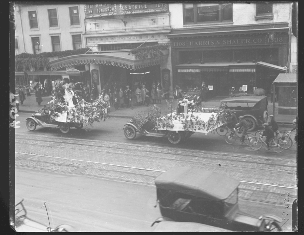 Schwarzes und weißes Foto eines Umzugs auf einer Stadtstraße mit Fahrzeugen einschließlich eines Autos und eines Fahrrads, Menschen auf dem Gehweg und Gebäude mit Fenstern und Namensschildern im Hintergrund.