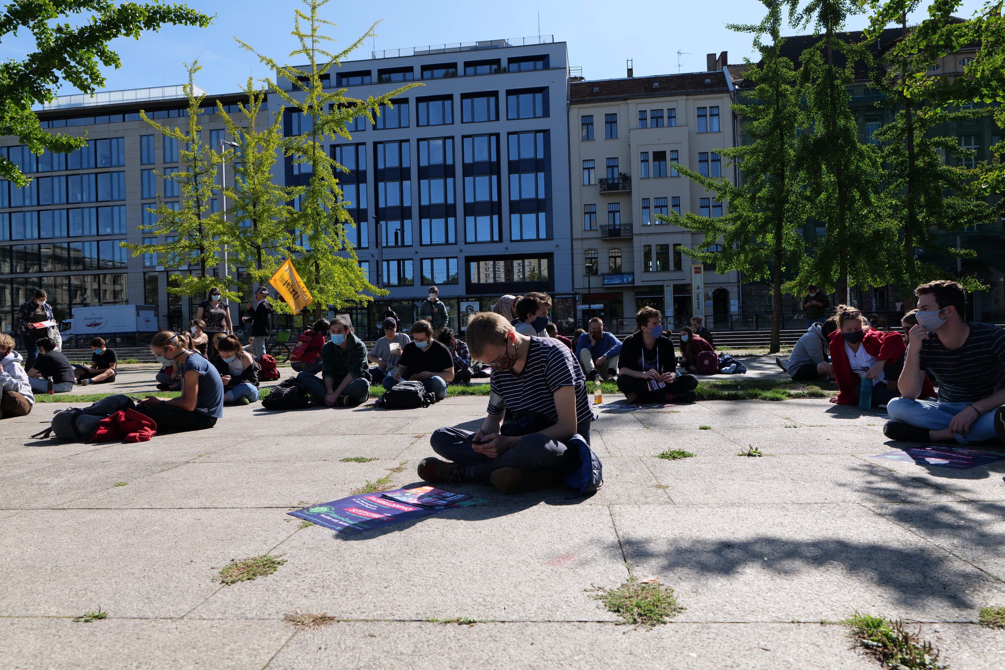 Menschen sitzen vor einem Gebäude auf dem Boden während einer Demonstration in Berlin, einige tragen Masken, mit verstreuten Taschen und Bäumen unter einem klaren blauen Himmel.