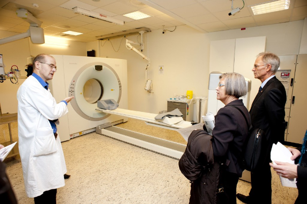 Arzt in weißer Uniform, der eine CT-Scan-Maschine zwei Personen in schwarzen Mänteln erklärt.