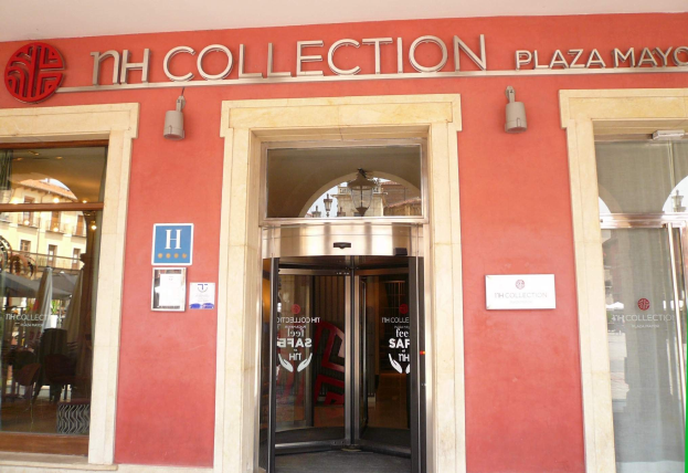 Eingang zum NH Collection Plaza Mayor Gebäude mit einer Glastür, flankiert von Fenstern, und einer beleuchteten Decke darüber; drinnen sind Möbel und Schirme sichtbar.