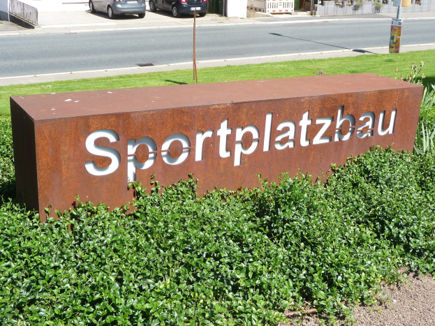 Sign reading "Sportplatzbau" in front of a building, with lush green grass and plants, and vehicles, buildings, and other objects in the background.