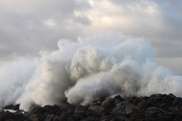 Große Welle, die gegen die Felsen am Strand schlägt, mit Wasser, das an den Strand schwappt, und bewölktem Himmel im Hintergrund.