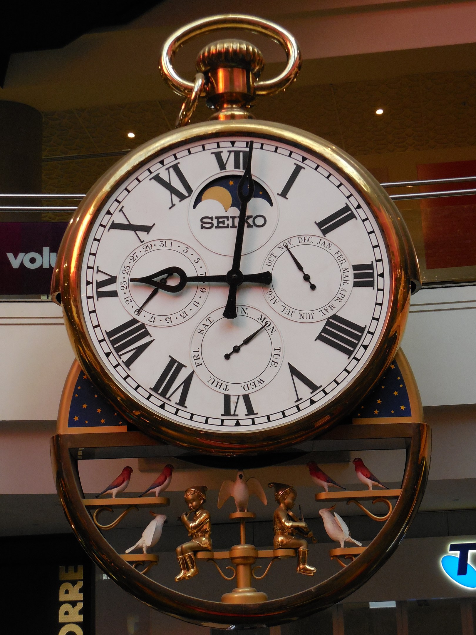 Goldene Taschenuhr, die an der Seite eines Gebäudes hängt, Zifferblatt mit römischen Zahlen und Text, Hintergrund zeigt eine Tafel mit Text und Deckenbeleuchtung.