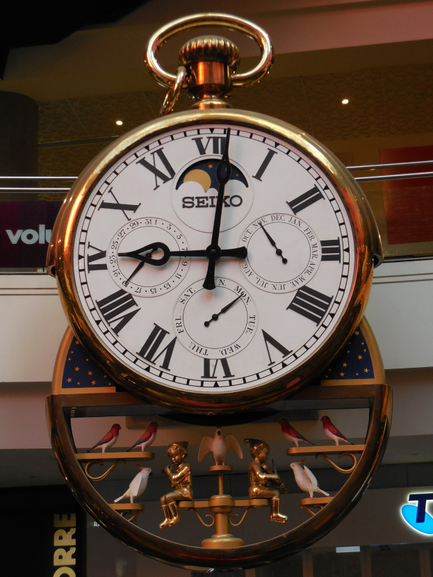 Goldene Taschenuhr, die an der Seite eines Gebäudes hängt, Zifferblatt mit römischen Zahlen und Text, Hintergrund zeigt eine Tafel mit Text und Deckenbeleuchtung.