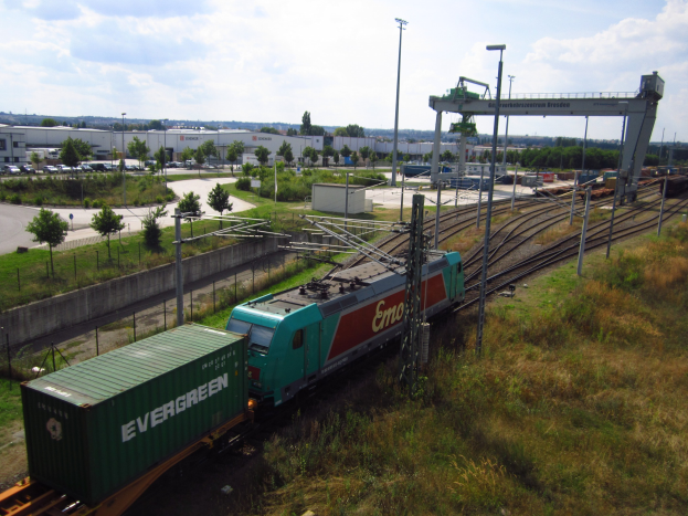 Güterzug fährt auf Schienen neben einem grünen Feld mit Strommasten, Kabeln, Bäumen, Gebäuden, Fahrzeugen und einem bewölkten Himmel im Hintergrund.