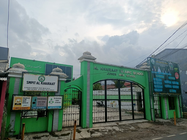 Eingang zur Al Khairat Islamischen Schule für Junioren mit einem grünen Tor, Beschilderung, umgebenen Pfählen und Hintergrundgebäuden, Bäumen und Himmel.