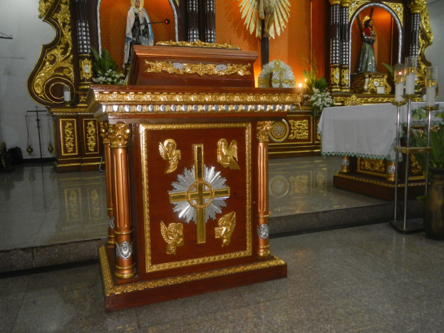 Altar in einer Kirche mit einem Kreuz, umgeben von Statuen, Blumenbouquets, Kerzen mit Ständern, einem tischdecke-überzogenen Tisch und Wandlichtern, die eine feierliche Atmosphäre schaffen.