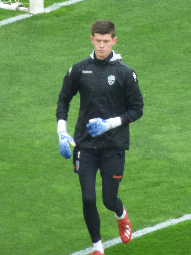 Ein professioneller Fußballspieler in schwarzer Jacke und blauen Fußballhandschuhen auf einem Fußballfeld mit einem Tor im Hintergrund.