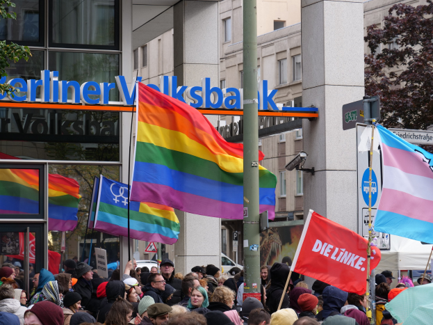 Eine große Gruppe von Menschen, die vor einem Gebäude stehen und Fahnen und Plakate mit Texten halten, bei einer Christopher Street Day Parade in Berlin, mit Bäumen zu beiden Seiten und Gebäuden im Hintergrund.