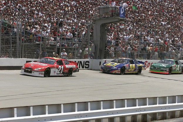 Drei NASCAR-Autos auf der Rennstrecke mit einer Zuschauermenge im Hintergrund, umgeben von Absperrungen und Rennstreckenschildern.