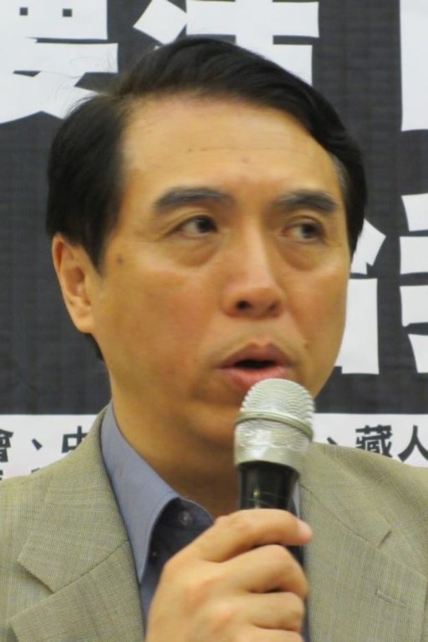 A man in a navy blue suit, white shirt, and patterned tie speaks into a microphone with a serious expression, standing in front of a banner with text.