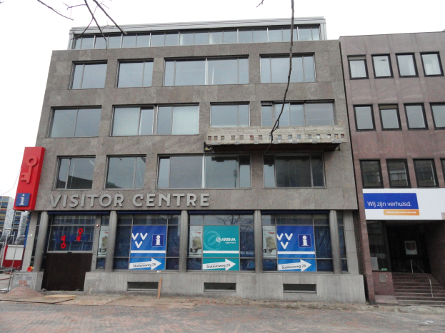 Großes Gebäude mit einer "Besucherzentrum"-Aufschrift, umgeben von anderen Gebäuden, Bäumen und einem klaren blauen Himmel.