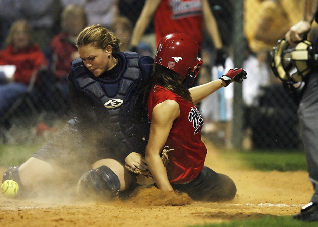 Softball-Spielerin rutscht mit Helm und Handschuhen auf Home Plate, umgeben von Maschendrahtzaun mit Zuschauern im Hintergrund auf einem Rasenfeld.