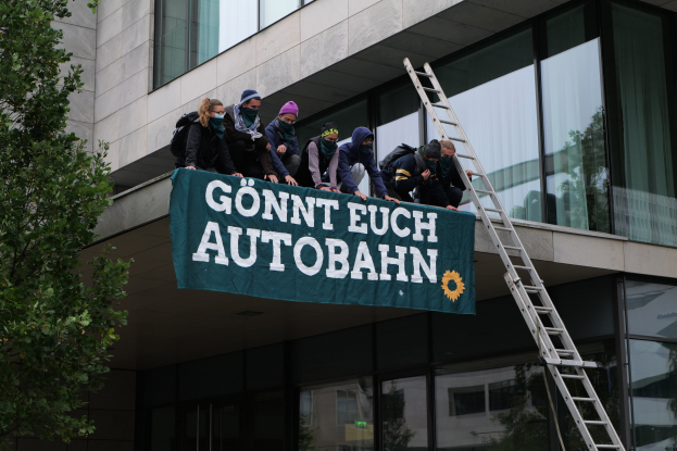 Gruppe von Menschen auf einem Gebäude, die eine Fahne halten, mit einem Baum links und Glasfenstern im Hintergrund, die scheinbar für die deutsche Regierung protestieren.