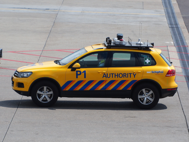 Gelbes Polizeifahrzeug mit 'TAXI'-Text an der Seite auf dem Rollfeld eines Flughafens geparkt, mit einer Person im Inneren und einem weiteren Fahrzeug teilweise links sichtbar.