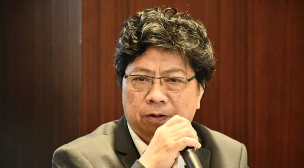 Mann in Anzug und Krawatte mit Mikrofon, der eine hitzige Diskussion zu führen scheint, vor einem Hintergrund aus Holzlatten.