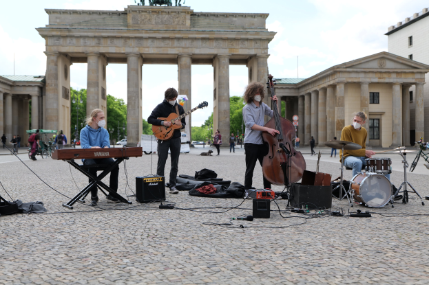 Group of people performing music outdoors with a guitarist seated, another musician playing an instrument, and a masked individual, in front of the Brandenburg Gate with scattered equipment and onlookers in the background.