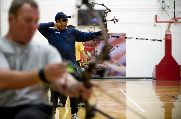 Männer beim Hallenbogenschießen, jeder mit einem Bogen und einem Pfeil in der Hand, mit einem Basketballkorb im Hintergrund.