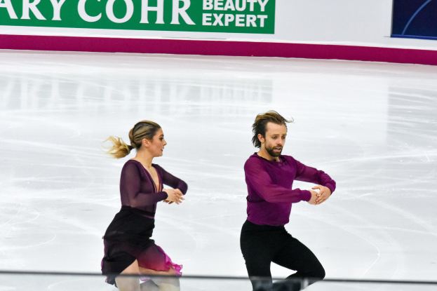 Zwei Eiskunstläufer, ein Mann und eine Frau, führen synchronisierte Bewegungen auf einer Eisbahn durch, im Hintergrund ist eine Tafel mit "Tessa Virtue und Scott Moir" zu sehen.