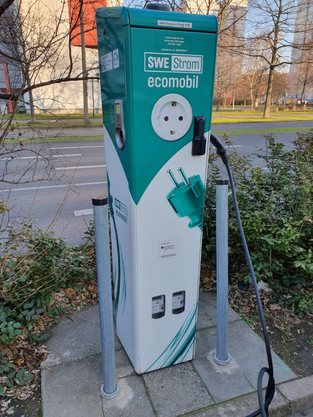 Elektrische Fahrzeug-Ladestation mit einer grünen und weißen Box und einem angebrachten Kabel, das an der Seite einer Straße mit Gebäuden, Bäumen und einem klaren blauen Himmel im Hintergrund steht.