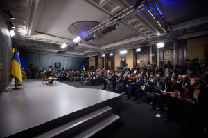 Eine große Gruppe von Menschen, die auf Stühlen vor einer Bühne mit einem zentralen Tisch sitzen, eine Flagge auf der linken Seite und Kameramänner auf der rechten Seite in einem Raum mit Türen und Deckenleuchten während einer Pressekonferenz in der Ukraine.