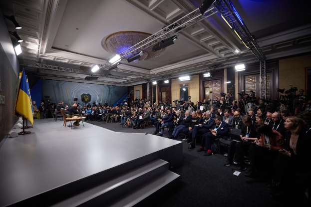 Eine große Gruppe von Menschen, die auf Stühlen vor einer Bühne mit einem zentralen Tisch sitzen, eine Flagge auf der linken Seite und Kameramänner auf der rechten Seite in einem Raum mit Türen und Deckenleuchten während einer Pressekonferenz in der Ukraine.
