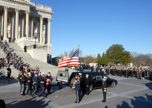 Beerdigungsprozession mit Menschen in Mützen und Fahnen vor dem U.S. Capitol Building, Fahrzeuge auf der Straße, Treppen mit Geländern, Laternenpfähle, Bäume und ein Himmel im Hintergrund.