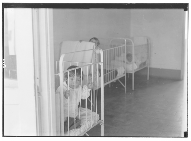 Schwarz-weiß-Foto eines Kindes in einem Krankenhauszimmer mit Feldbetten, Decken, Kissen, einer Tür auf der linken Seite, einer Wand im Hintergrund und einem sichtbaren Boden.