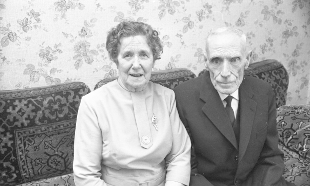 Zwei ältere Erwachsene, ein Mann in einem Anzug und eine Frau in einer unbestimmten Garderobe, sitzen auf einer Couch und schauen ernst nach vorne.