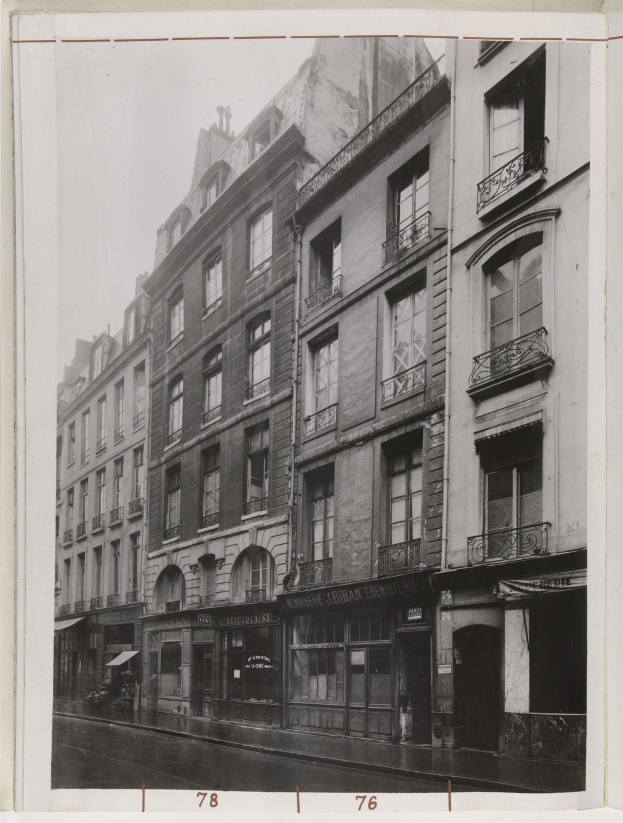 Schwarzes und weißes Foto einer Stadtstraße mit Gebäuden mit Fenstern, Geländern und Namensschildern sowie einer Straße unten.