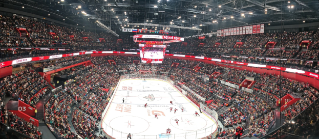 Eine große Indoor-Hockey-Arena mit Zuschauern in den Rängen, heller Deckenbeleuchtung und einem zentralen Scoreboard-Bildschirm.