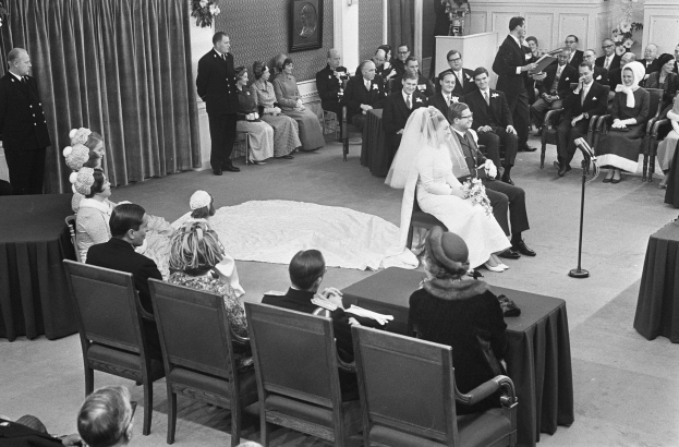 Schwarze und weiße Fotografie einer Hochzeitszeremonie mit sitzenden und stehenden Menschen, einem Mann mit Mikrofon, einer Frau mit Blumen, einem Podium mit Mikrofon, Blumenschmuck, einem Vorhang, einem gerahmten Foto an der Wand und einer Tür im Hintergrund.