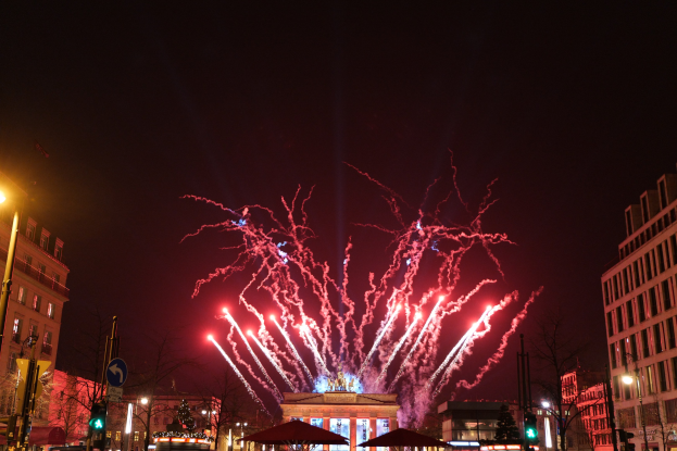 Eine belebte Stadtstraße während einer Silvesterfeier in Berlin mit Menschenmassen, Fahrzeugen, Gebäuden und festlichen Feuerwerken, die den Nachthimmel erhellen.
