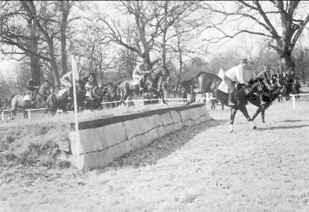 Black and white photo of people riding horses over a grassy field with a wall and poles in the foreground, trees and sky in the background, and text at the bottom reading "Horse Jumping at the National Horse Show in Washington, D.C. USA."