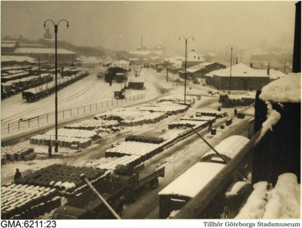 Schwarzes und weißes Foto einer schneebedeckten Stadtstraße mit Fahrzeugen, Polen, Laternen, Gebäuden, Bäumen, einem Zaun und Text am unteren Rand.