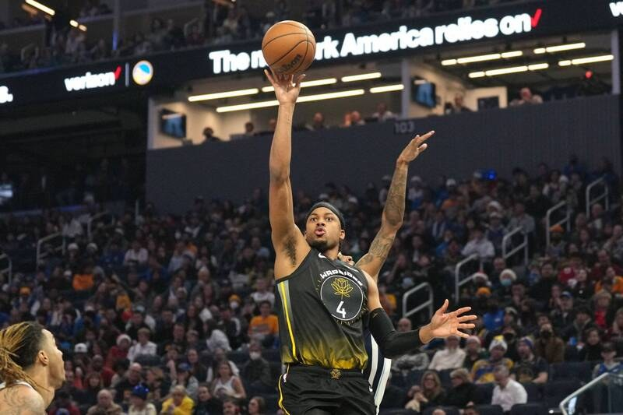 Ein Basketballspieler in einem schwarzen und gelben Trikot der Golden State Warriors schießt einen Ball in einer hell erleuchteten Arena vor einer Menge von Zuschauern.