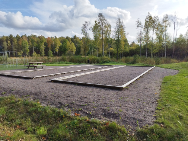 Ein grasbewachsener Platz mit einer Picknick-Tafel, umgeben von Bäumen unter einem klaren blauen Himmel, mit Spielplatz-Geräten einschließlich Stangen und einer Bank auf der linken Seite.